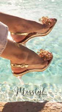 Load image into Gallery viewer, A pair of gold-colored flat sandals with ankle straps, displayed in a manner that suggests they are being worn by a person sitting by a poolside.